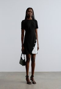 Vestido preto até ao joelho com mangas curtas, apresentando um painel branco diagonal e detalhes de botões na lateral. Combinado com uma pequena mala preta e saltos altos.