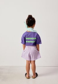 Enfant debout, dos à la caméra, portant un t-shirt violet "American Vintage", un short violet clair, des sandales noires et un foulard bleu clair autour du cou.