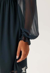 Mörk teal, genomskinlig klänning med långa, böljande ärmar. Ärmsluten är krusiga och visar upp ett lätt, texturerat tyg.