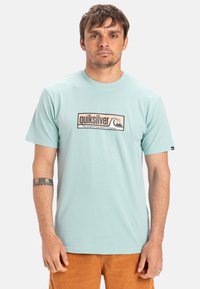 Tyrkysové tričko s krátkým rukávem a logem Quiksilver v obdélníkovém designu; měkká tkanina a kulatý výstřih, spárované s béžovými kordovým šortkami.