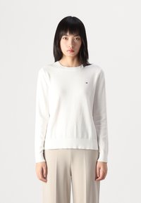 Maglione bianco a maniche lunghe con scollatura rotonda, tessuto testurizzato e un piccolo logo sul lato sinistro del petto. Abbinarlo a pantaloni beige chiaro.