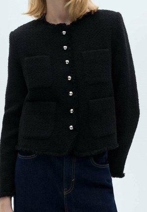 Cardigan noir texturé boutonné avec six boutons argentés et quatre poches avant, porté avec un jean bleu foncé.