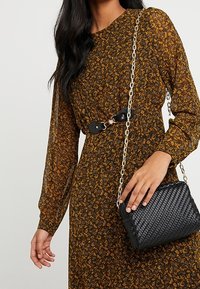 Sac à main tissé noir avec une bandoulière en chaîne, associé à une robe marron à motifs floraux avec des manches longues et une taille cintrée.