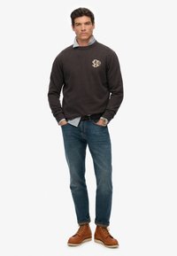 Felpa grigio scuro con scollatura a girocollo e logo sul petto, indossata sopra una camicia a scacchi blu chiaro. Abbinata a jeans blu e stivali marroni.