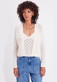 Cárdigan de encaje de ganchillo en color crema, diseño de frente abierto, patrón texturizado, usado sobre una blusa ligera, combinado con pantalones de mezclilla de cintura alta.