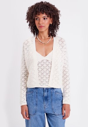 Häkeltop in Creme, offenes Frontdesign, strukturierter Muster, getragen über einem leichten Top, kombiniert mit hochtaillierten Jeans.