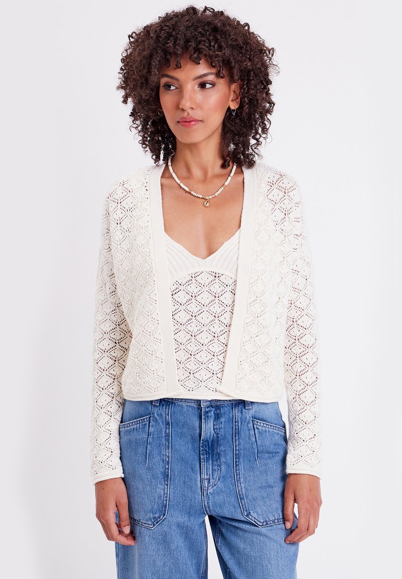 Cárdigan de encaje de ganchillo en color crema, diseño de frente abierto, patrón texturizado, usado sobre una blusa ligera, combinado con pantalones de mezclilla de cintura alta.
