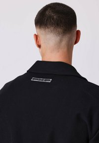 Giacca nera con colletto e una patch con logo sul retro. Presenta un tessuto testurizzato e spalle strutturate.