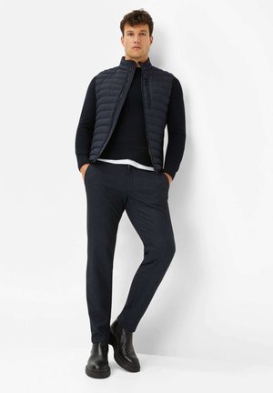 Marineblauer gesteppter Weste über einem schwarzen Oberteil, kombiniert mit dunkel gemusterten Hosen und schwarzen Stiefeletten, die ein minimalistisches Design und funktionale Reißverschlüsse aufweist.