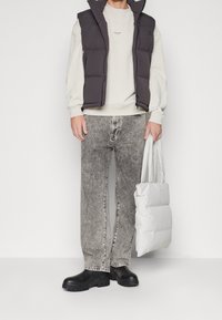 Chaleco de plumas en gris oscuro sobre un sudadero claro, combinado con jeans gris claros y botas negras gruesas. Un bolso blanco doblado completa el conjunto.