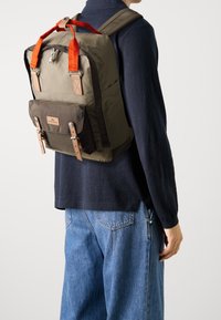 Khaki backpack with red straps, featuring a front pocket, leather accents, and metal hardware. Textured fabric with a structured shape.