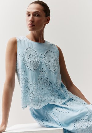 Femme portant un haut sans manches en dentelle œillet bleu clair et une jupe assortie, assise de côté avec une expression neutre sur un fond uni.