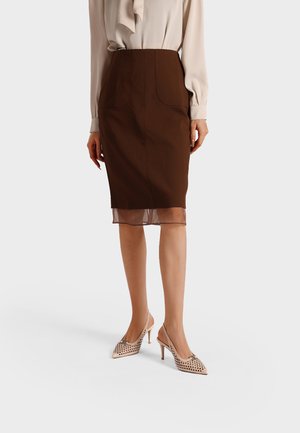 Donna che indossa una gonna marrone al ginocchio con bordo trasparente, camicetta beige e décolleté slingback beige con punta appuntita e ritagli circolari.