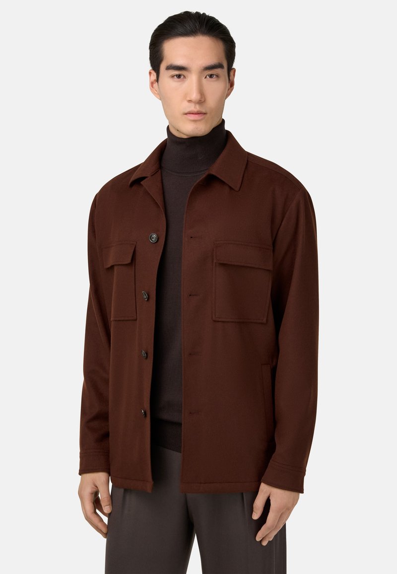 Braune, weiche Stoffjacke mit kragenlosem Ausschnitt, Knopfverschluss und zwei Brusttaschen, getragen über einem dunklen Rollkragenpullover. Minimalistisches Design.