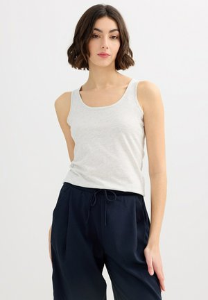 Joven mujer con cabello corto y oscuro, vistiendo una camiseta sin mangas gris claro y pantalones de cordón azul marino oscuro, de pie frente a un fondo blanco.