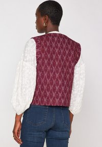 Donna con i capelli corti che indossa una blusa bianca con ricami e maniche a sbuffo, un gilet senza maniche rosso scuro effetto testo, e jeans blu, mostrata di spalle.