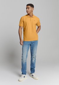 Geel polo shirt, korte mouwen, met een kraag en klein embleem. Gemaakt in combinatie met lichtblauwe jeans en witte sneakers, tegen een neutrale achtergrond.