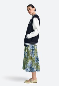 Fekete-fehér egyetemi dzseki csíkos mandzsettával, zöld és kék árnyalatú virágos szoknyával, és világos színű balerinacipővel párosítva.