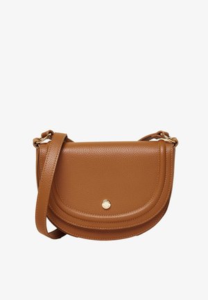 Kvinde iført en striped knaptrøje, hvide bukser, hvide loafers og en brun skuldertaske, stående sidelæns mod en beige baggrund.