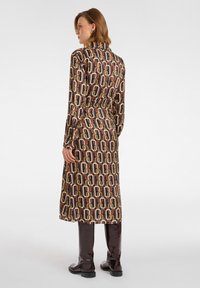 Sukienka z długim rękawem w wzorzystym materiale, z geometrycznymi kształtami w odcieniach brązu, pomarańczu i kremu. Wyposażona w dopasowaną talię i detale przy kołnierzu.