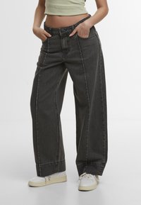 Personne portant un jean en denim noir ample et des baskets blanches, debout les mains dans les poches devant un fond uni.