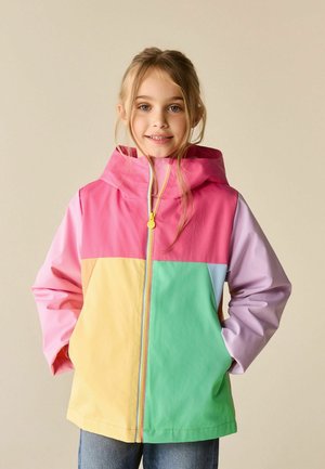 Young girl smiling, wearing a multicolored hooded jacket with pink, yellow, green, and purple panels, hands in pockets, light beige background.