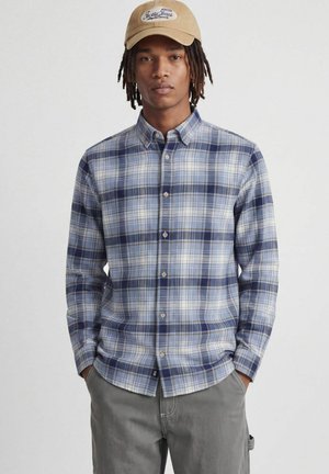 Jeune homme avec des dreadlocks portant une casquette beige, une chemise à carreaux bleu et blanc boutonnée, et un pantalon gris, les mains dans les poches.