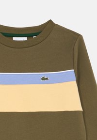 Πράσινο βαμβακερό πουλόβερ με οριζόντιες ρίγες σε ανοιχτό μπλε και κίτρινο, με ένα μικρό κεντημένο λογότυπο κροκοδείλου στο στήθος.