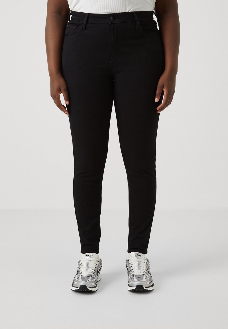 Jeans negros ajustados fabricados con una tela elástica, con un diseño clásico de cinco bolsillos, cierre de cremallera y longitud recta hasta el tobillo. Combinados con zapatillas plateadas.