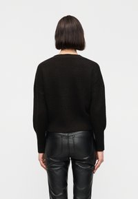 Γυναίκα με κοντά σκούρα μ αλλιά φοράει μαύρο πλεκτό πουλόβερ και μαύρο δερμάτινο παντελόνι, γυρισμένη προς τα πίσω μπροστά σε μονόχρωμο λευκό φόντο.