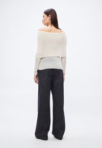 Svetlobéžový top s odhalenými ramenami v kombinácii so sivou spodnou vrstvou a voľnými tmavými denimovými širokými nohavicami, ktoré zdôrazňujú uvoľnený strih a mäkké textúry.