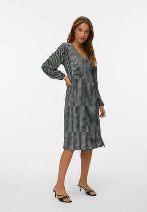 Femme posant dans une robe portefeuille à manches longues à carreaux noir et blanc, de longueur midi, avec des sandales à talons noirs.