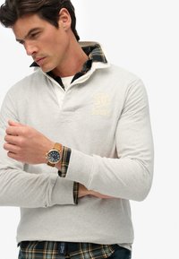 Lys grå rugby-style sweatshirt med en hvid krave og en ternet skjorte under. Har et broderet logo og et træur på håndleddet.