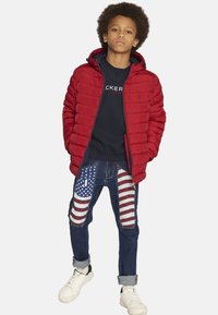 Giacca puffer rossa con zip e cappuccio, felpa blu navy, jeans in denim strappati con toppe a bandiera americana e sneakers bianche.