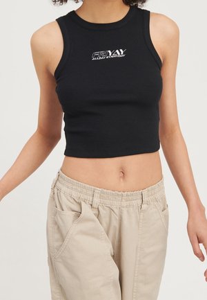 Frau trägt ein schwarzes, kurzes Tanktop mit dem Schriftzug "FRIYAY ALL DAY EVERYDAY" und beige Hosen mit elastischem Bund vor einfachem Hintergrund.