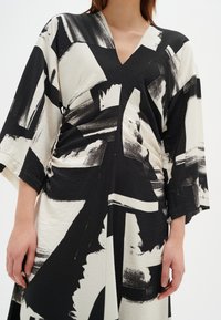 Mustavalkoinen mekko, jossa on abstrakteja siveltimen vetoja, V-pääntie ja 3/4 hihat pehmeässä, teksturoidussa kankaassa.