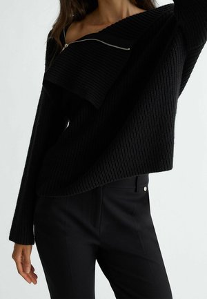 Femme portant un pull en maille côtelée noir avec une fermeture éclair asymétrique et un pantalon noir moulant sur un fond uni.