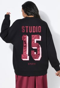 Zwart oversized sweatshirt met de tekst "STUDIO 15" in gestructureerde bordeauxrode letters met glanzende accenten. Soepele stof met geribbelde manchetten.