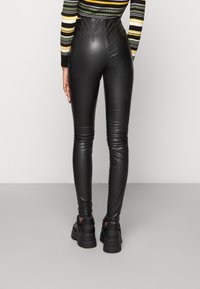 Kvinna som bär svarta högmidjade leggings i konstläder, svarta platåskor och en långärmad randig topp, stående med ryggen mot kameran.