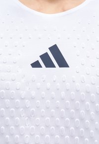 Gros plan d’un maillot de sport blanc texturé avec des points en relief et un logo Adidas noir centré sur la poitrine.