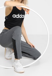 Czarny krótki top z białym logo „adidas”, szare spodnie z czarnymi bocznymi akcentami oraz białe buty sportowe, pozujący z okrągłą obręczą.