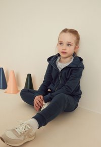 Hanorac și pantaloni de trening moi de culoare albastru marin, tricou gri dedesubt, adidași albi cu accente deschise, copil așezat pe podea bej cu conuri colorate în apropiere.