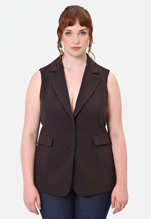 Donna con capelli rossi che indossa un blazer nero senza maniche con due bottoni e jeans blu scuro, in piedi davanti a uno sfondo chiaro e semplice.