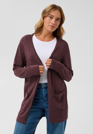 Femme aux cheveux châtain clair portant un cardigan marron par-dessus un haut blanc et un jean bleu, sur un fond clair uni.