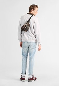 Kamouflage midjeväska bärs över en grå sweatshirt, ihop med ljusblå jeans och vinröda sneakers. Detaljer av avslappnad urban outfit synliga.