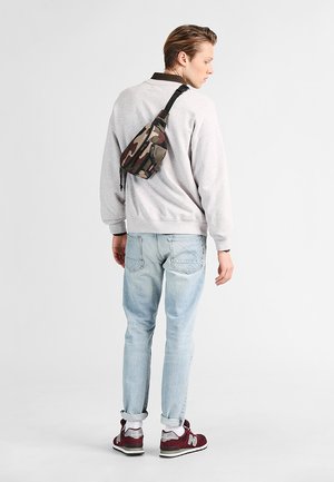 Kangashinti, joka on käytössä harmaan hupparin päällä, yhdistettynä vaaleansinisiin farkkuihin ja tummanpunaisiin tennareihin. Rennon urbaanin vaatetuksen yksityiskohdat näkyvät.