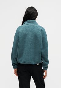 Άτομο με μακριά σκούρα μαλλιά που φοράει γαλάζιο φλις μπουφάν και μαύρο παντελόνι, γυρισμένο προς τα πίσω μπροστά σε απλό λευκό φόντο.