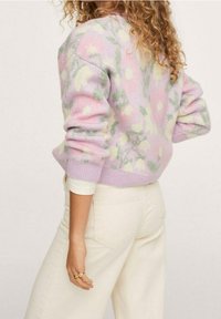 Pull rose pastel à imprimé floral vert et jaune. Présente des poignets et un ourlet côtelés, fabriqué en tissu tricoté doux, porté avec un jean blanc.