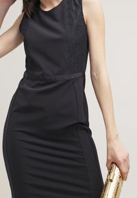 Svart klänning med en spetspanel på vänster sida, figurnära silhuett och en slät textur; matchad med en metallisk guldväska.