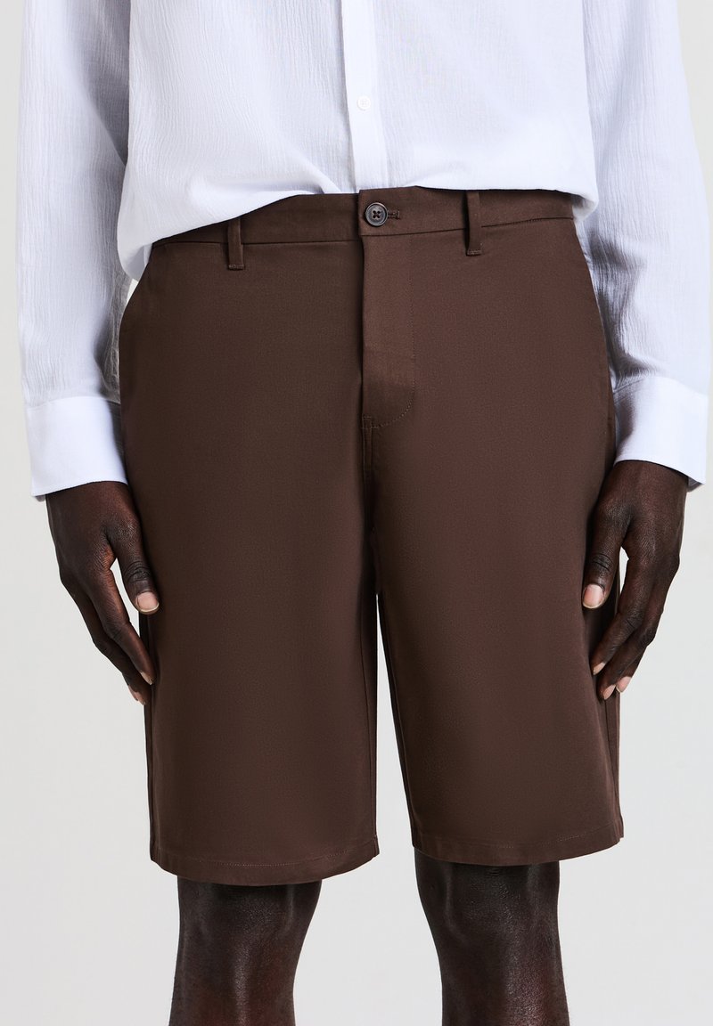 Homme portant un short marron ajusté et une chemise blanche à manches longues, se tenant les mains détendues sur les côtés sur un fond uni.